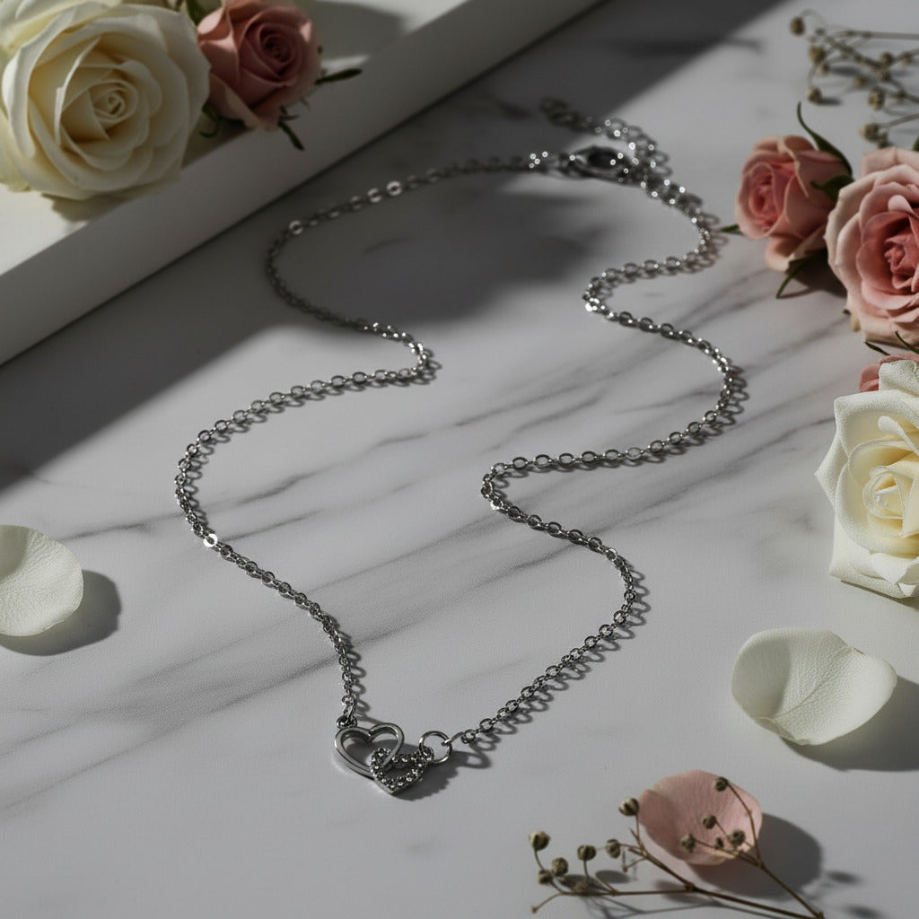 A delicate silver chain necklace with a double heart pendant, displayed on a soft beige satin fabric background.