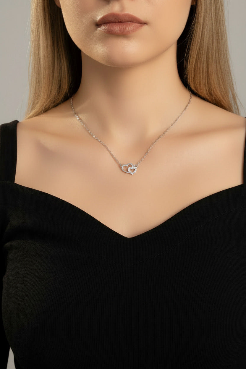 A close-up of a model's neck and chest wearing a delicate silver chain necklace with a double interlocking heart pendant.